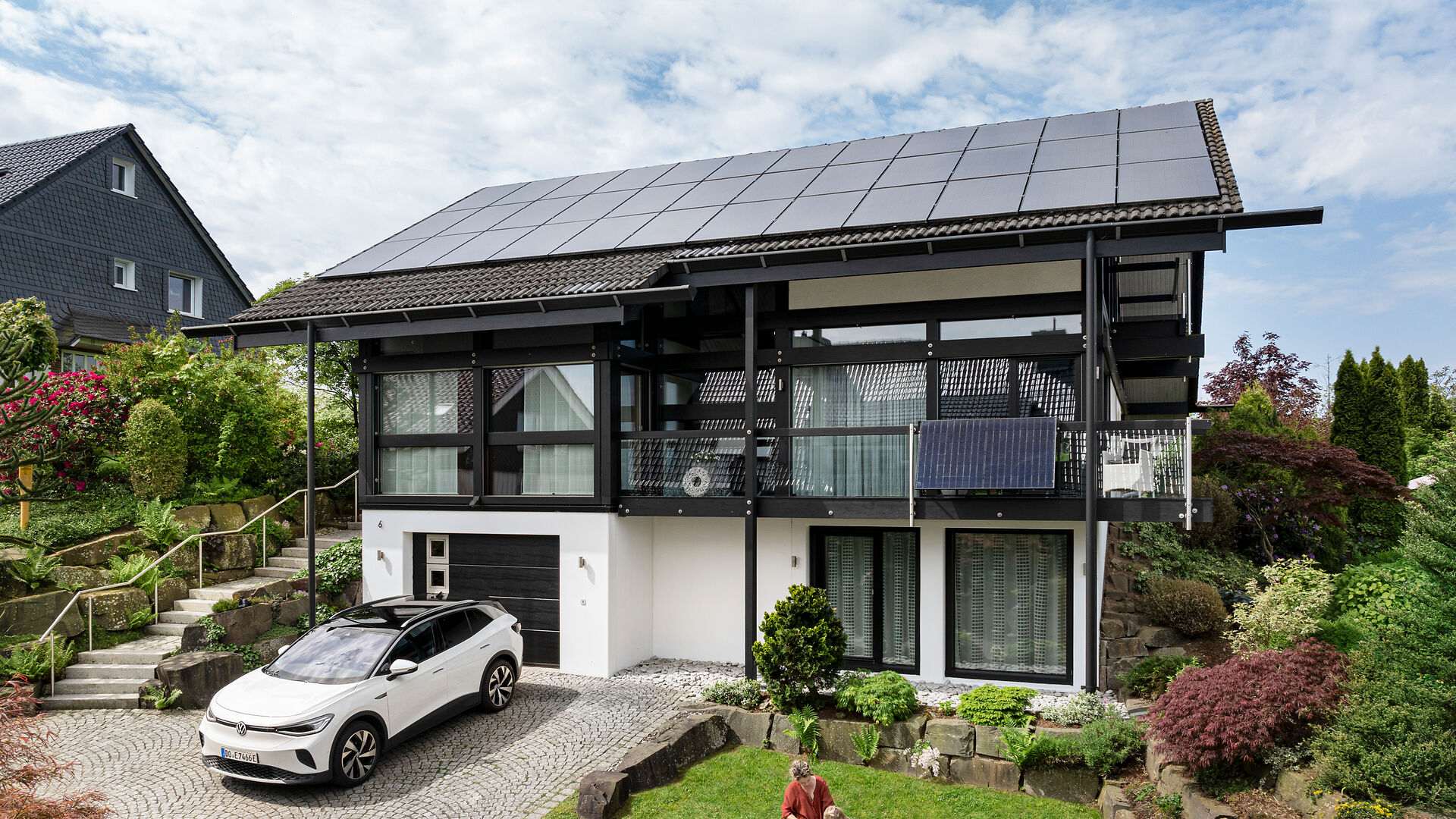 Modernes Haus mit Solardach und E-Auto in Einfahrt, umgeben von Garten und Nachbarhaus