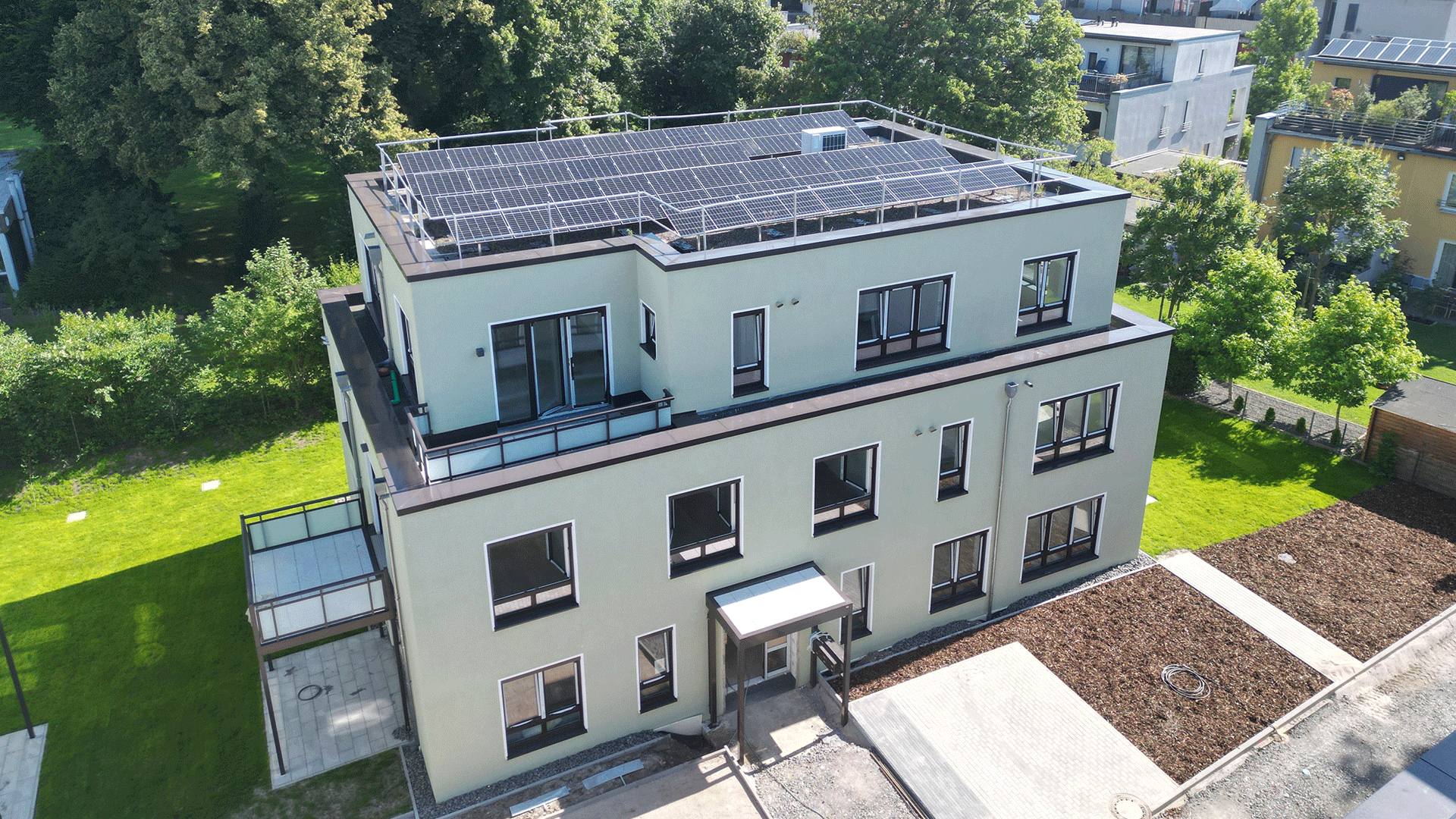 Modernes Gebäude mit Flachdach und Solarpanels, umgeben von Grünflächen und Bäumen.