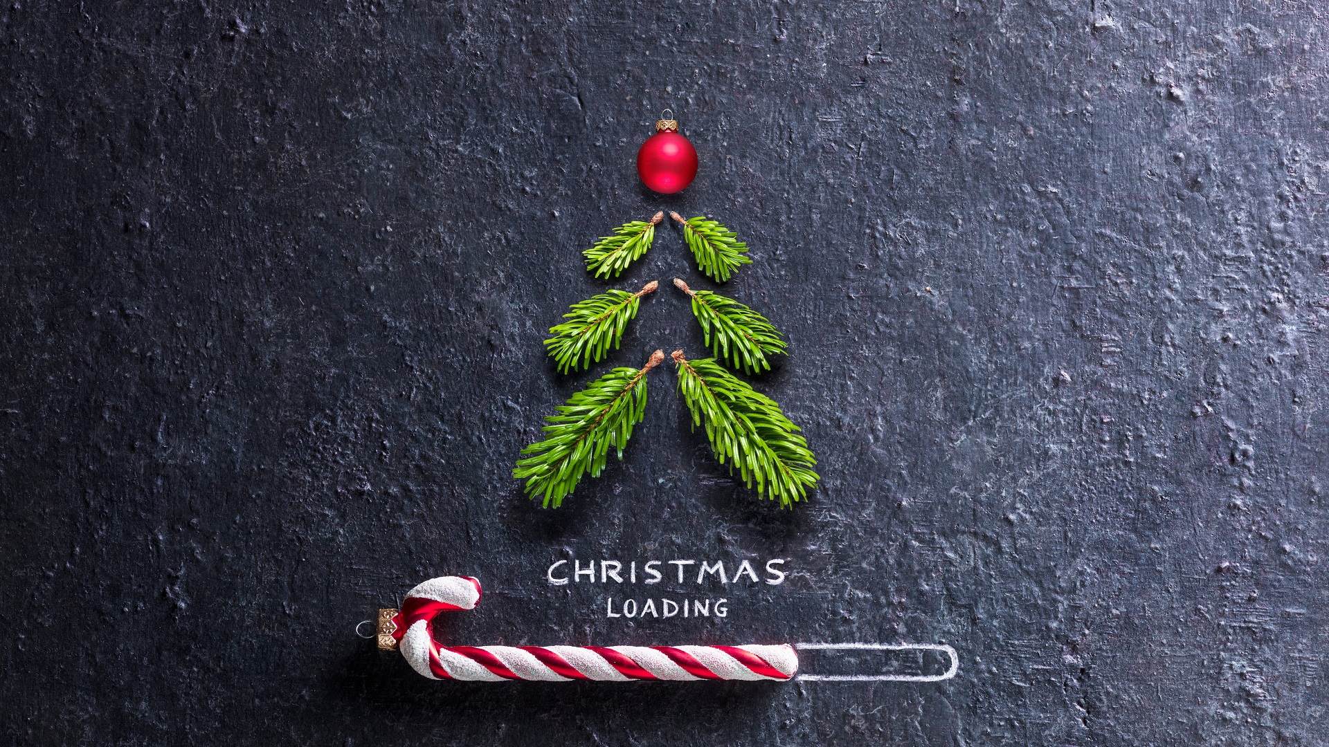 Weihnachtsbaum aus kleinen Zweigen und roter Kugel auf dunklem Hintergrund, darunter "CHRISTMAS LOADING" mit Zuckerstange im Ladebalken.