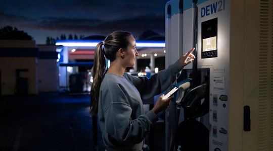 Ladestation Eine Frau an einer beleuchteten Ladestation für Elektrofahrzeuge des Unternehmens "DEW21" zeigt auf den Bildschirm der Station. Es ist dunkel, und im Hintergrund ist eine Tankstelle zu sehen.