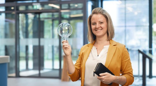 Stromkennzeichnung Person hält eine große Glühbirne und eine schwarze Geldbörse, trägt einen gelben Blazer.