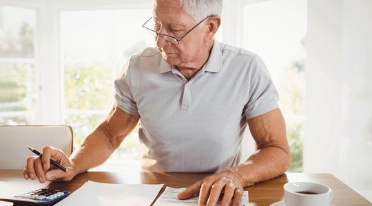 Berechnung der Kosten Ältere Person am Tisch mit Papieren, Stift und Taschenrechner, im Hintergrund helle Fenster.